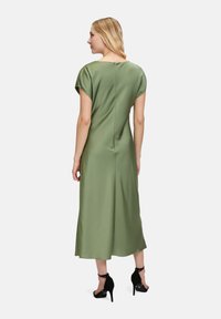 Robe verte olive, à mi-mollet, avec des manches courtes, un col rond et une fermeture éclair au dos. Portée avec des chaussures à talons hauts noires.