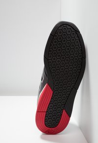 Černá podrážka sportovní obuvi s texturovaným vzorem včelí plástve. Obsahuje červené akcenty na patě a zakřivený boční design pro lepší přilnavost.