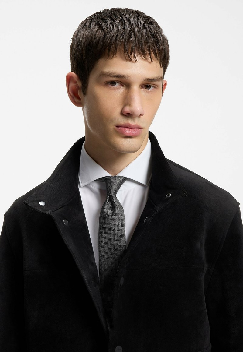 Jeune homme aux cheveux courts et foncés, portant un manteau noir sur une chemise blanche et une cravate grise à motifs, sur un fond blanc uni.