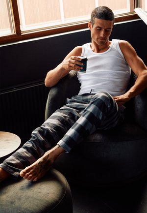 Grau und schwarze karierte Schlafanzughose, weißes Tanktop, sitzt auf einem runden, plüschigen Stuhl. Hält eine kleine, türkisfarbene Kaffeetasse. Tattoo am Fuß sichtbar.