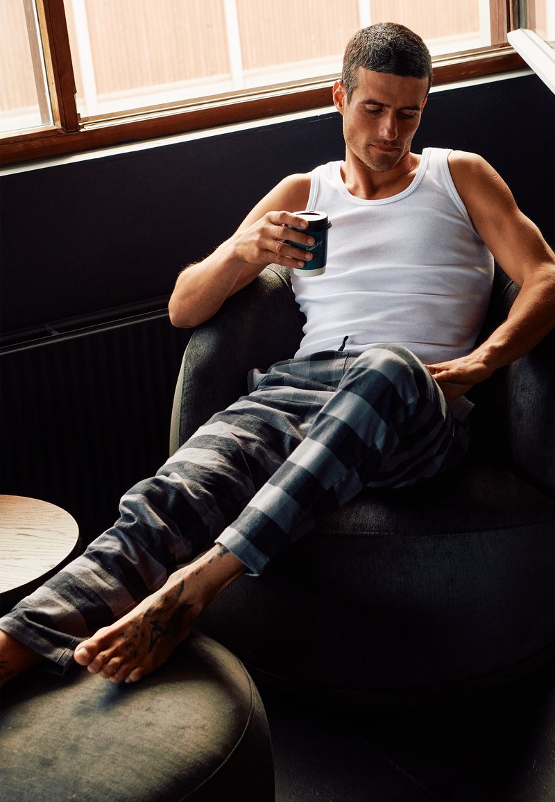 Grå og sort ternede pyjamasbukser, hvid tank top, siddende i en rund, blød stol. Holder en lille, teal kaffekop. Tatovering synlig på foden.