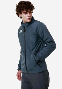 Giacca zip-up grigio-blu con colletto alto, frontale imbottito, tasche laterali e logo sul petto sinistro; realizzata in tessuto morbido e liscio.