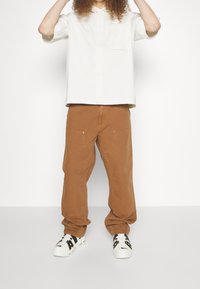 Lätt beige skjorta med krage och ficka. Löst sittande bruna byxor med böjda fickor. Vita sneakers med svarta detaljer.