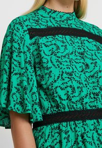 Vestido verde con estampado floral negro, con mangas cortas acampanadas, un escote alto y un detalle de encaje negro en la cintura.