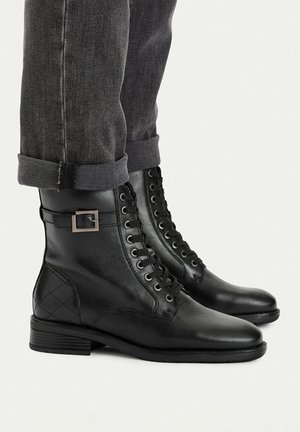 Černé kožené kotníkové boty s tkaničkami a boční přezkou, nošené s ohrnutými tmavě šedými džínami na světlém pozadí.
