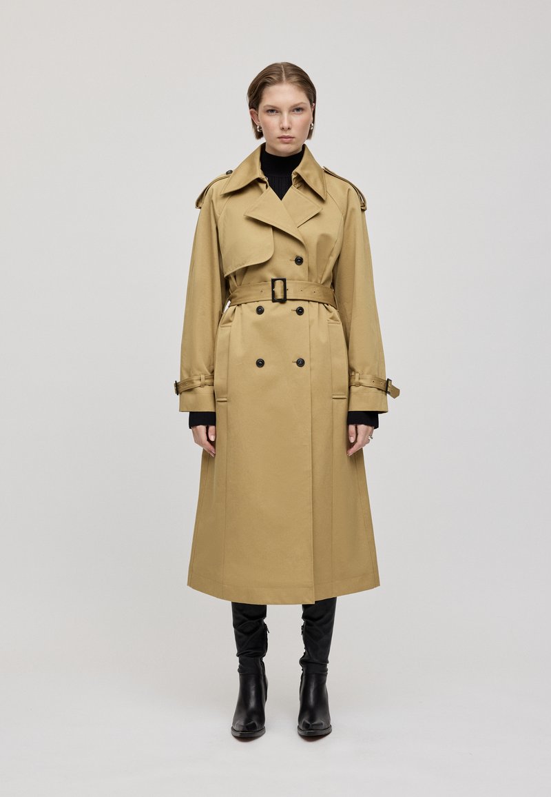 Cappotto trench color khaki con vita regolata da una cintura, frontale doppiopetto e spalline. Presenta maniche lunghe e taglio dritto, abbinato a stivali neri.