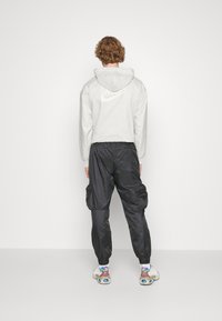 Person med lockigt hår som vänder bort ansiktet, klädd i en vit Nike-hoodie, svarta cargo-byxor, vita strumpor och färgglada sneakers mot en enfärgad bakgrund.