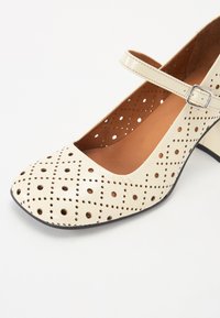 Pantofi din piele perforată de culoare crem, cu vârf rotund, curea cu cataramă argintie și toc mic pătrat. Interiorul este de culoare bej. Model texturat pe suprafață.