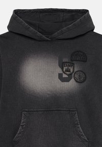Sweatshirt à capuche gris foncé avec une poche frontale, présentant trois patches noirs et un motif dégradé. Le matériau semble doux et texturé.