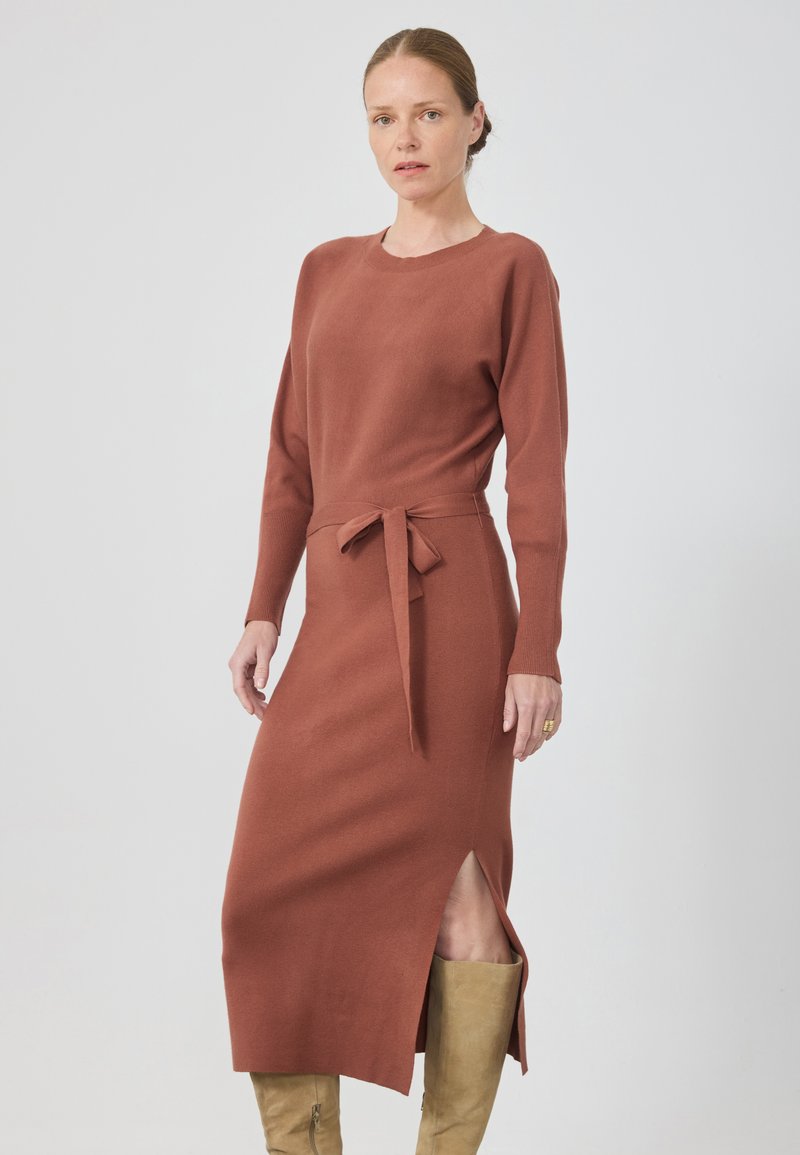 Robe en tricot de couleur rouille, avec des manches longues, un col rond, une taille ceinturée et une fente sur le côté. Texture lisse avec une silhouette ajustée.