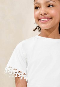 Hvid bomulds T-shirt med korte ærmer, der har dekorative blonde detaljer i kanten. Glat tekstur og klassisk rund halsudskæring.