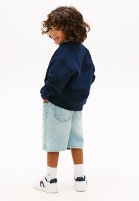 Χαμογελαστό παιδί με σγουρά μαλλιά στέκεται με τα χέρια στις τσέπες, φορώντας μπλε σκούρο φούτερ, ανοιχτόχρωμα τζιν σορτς, λευκές κάλτσες και λευκά αθλητικά παπούτσια.