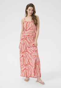 Robe maxi rouge avec motif de feuilles blanches, fines bretelles, taille froncée, fente sur le côté, portée avec des tongs blanches. Texture de tissu lisse.
