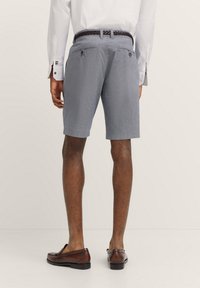 Herren Shorts in Marineblau mit Muster, geradem Schnitt, zwei Gesäßtaschen und einem kontrastierenden Gürtel. Kombiniert mit braunen Slippern und einem weißen Hemd.