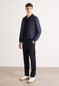 Navy bomber jacket with a front zipper, worn over a dark polo and paired with matching trousers and white sneakers with a tan sole.