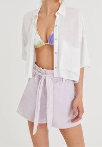 Femme portant une chemise blanche ample à boutons, un haut de bikini tie-dye, et un short taille haute violet pastel avec une ceinture en tissu assortie.
