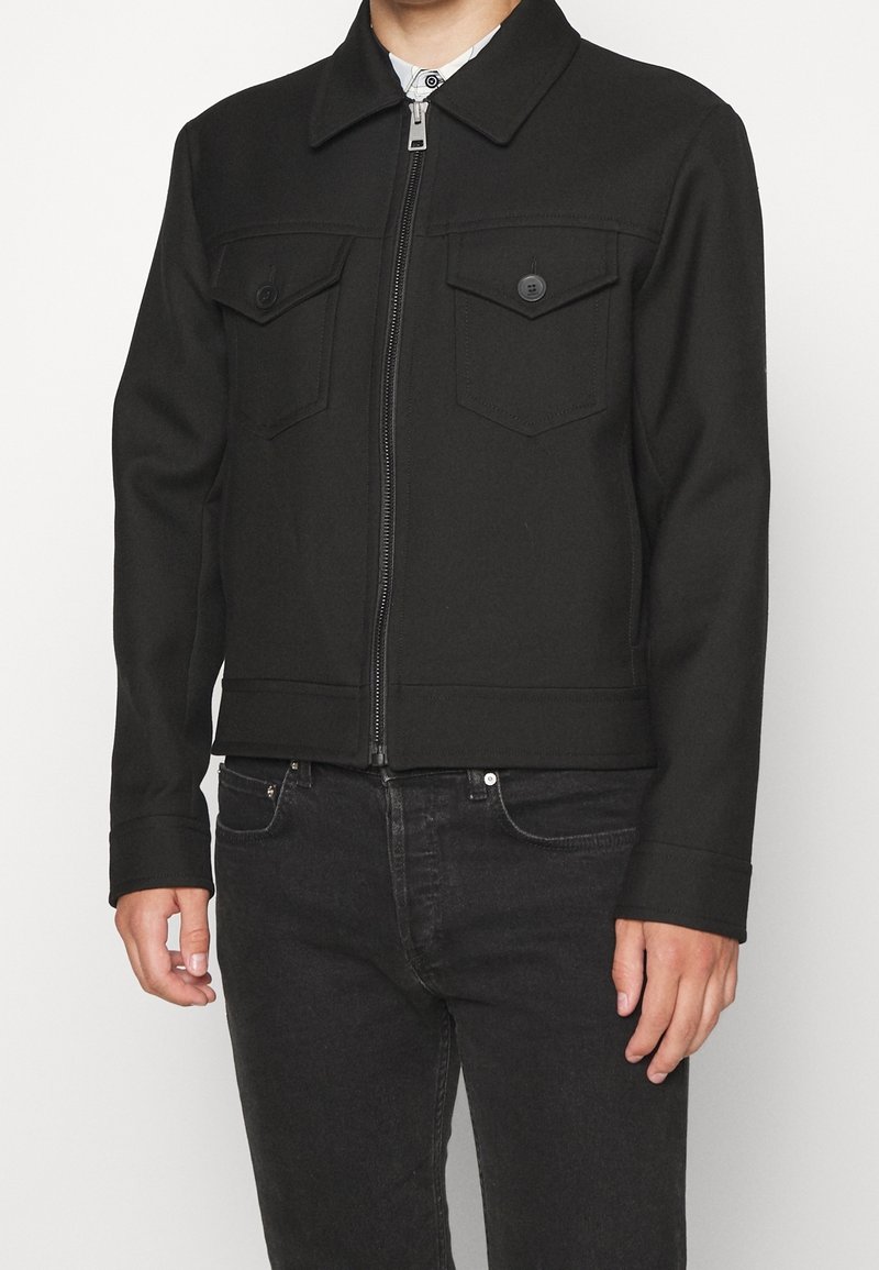 Homme portant une veste noire zippée avec poches poitrine boutonnées et un jean noir, debout devant un fond clair uni.
