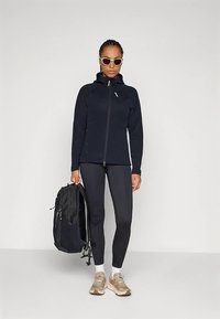 Schwarze Reißverschlussjacke, schwarze Leggings, beige Turnschuhe und ein schwarzer Rucksack. Sonnenbrillen unterstreichen den sportlichen, funktionalen Look.