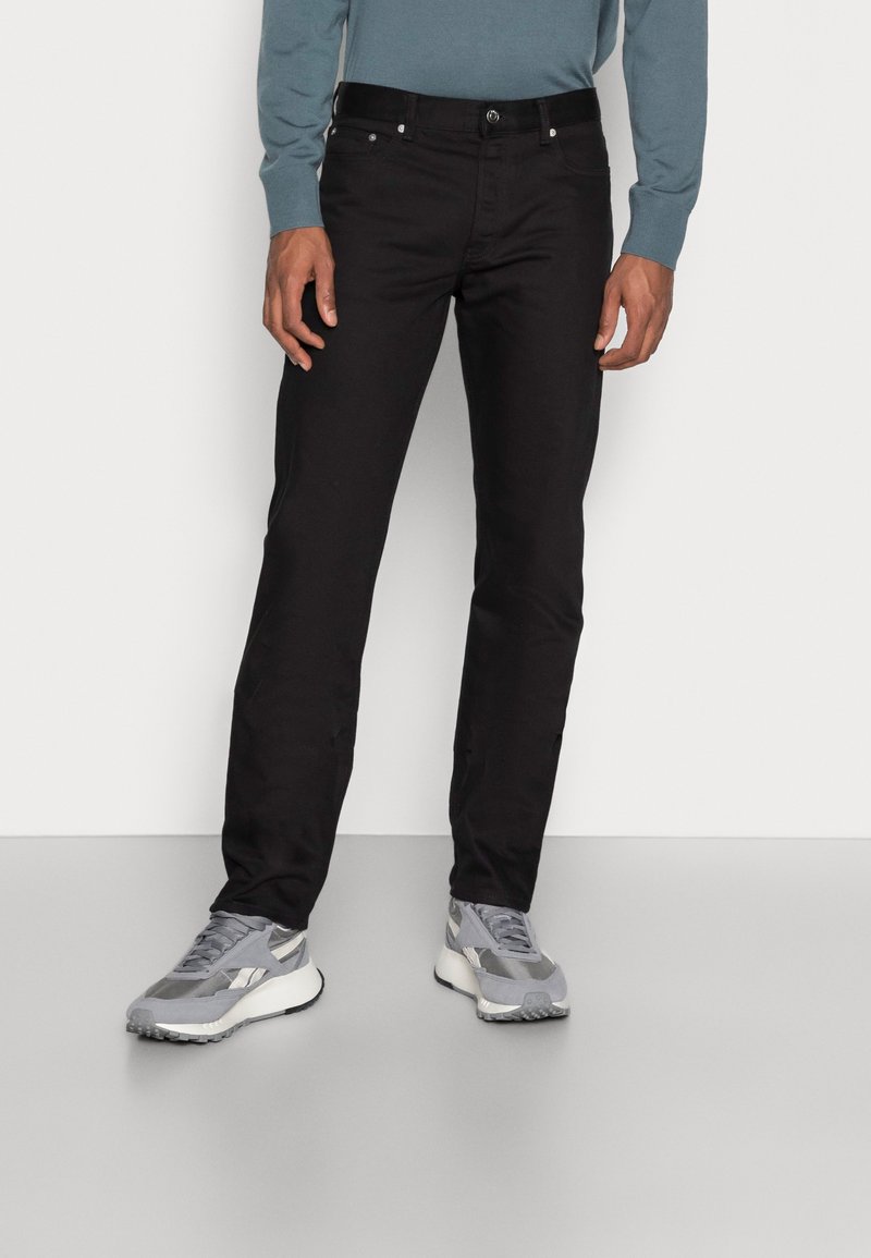 Homme portant un pantalon noir slim, des baskets grises et une chemise bleue à manches longues, debout devant un fond uni.