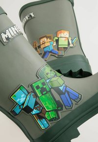 Zelené gumené dáždnikové topánky majú farebné nálepky postáv z Minecraftu, vrátane zombie a blokových postáv, s textúrovaným povrchom a pevným podošvou.
