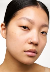 Gros plan d'un visage au teint clair et lisse, avec un anneau en argent au nez et trois gouttes de fond de teint sur la joue. Look de maquillage naturel.