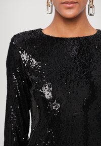 Top in paillettes nere con collo rotondo e maniche lunghe. Caratterizzato da una superficie testurizzata con dettagli brillanti e una silhouette aderente.