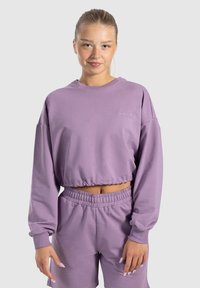 Felpa viola accorciata realizzata in morbido tessuto, con maniche lunghe, polsini a coste e un logo ricamato sul petto. Abbinata a pantaloni corti coordinati.