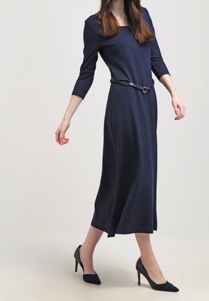 Woman in a navy blue midi dress with three-quarter sleeves, a thin belt, and black high-heeled shoes walking against a plain background.