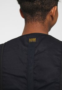 Vue de dos d'une personne portant un gilet en jean noir G-Star RAW avec une étiquette logo jaune en haut au centre du dos.