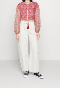 Donna che indossa un blusa floreale rossa trasparente con maniche lunghe, pantaloni cargo bianchi a gamba larga e sneakers nere basse su uno sfondo semplice.