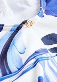 Gros plan sur un tissu bleu et blanc avec un imprimé floral abstrait et une petite boucle ronde dorée attachée à la couture.