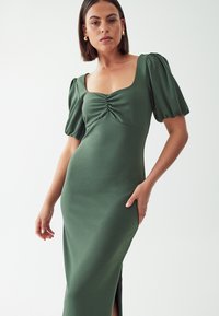 Olivno zeleno oprijeto obleko z napihnjenimi rokavi, kvadratnim izrezom in nabrano podrobnostjo na prsih, ki ima stranski razporek za dodatno gibanje.