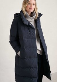 Abrigo acolchado largo de color azul marino oscuro con capucha, diseño acolchado, cierre de cremallera y bolsillos laterales, usado sobre un suéter gris claro.