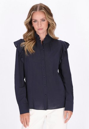 Hullámos szőke hajú nő sötét tengerészkék, vállán fodrokkal díszített hosszú ujjú blúzt és fehér nadrágot visel egyszínű háttér előtt.