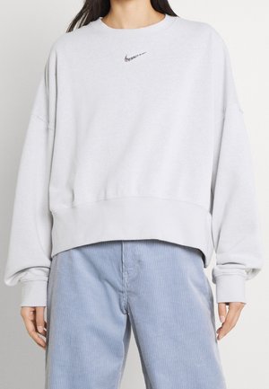 Persoon draagt een wit Nike sweatshirt met een klein logo op de borst en losse lichtblauwe corduroy broek.