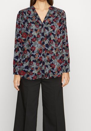 Vrouw draagt een blouse met lange mouwen en knopen, met een rood, paars en groen bloemenpatroon, gecombineerd met zwarte broek met wijde pijpen.