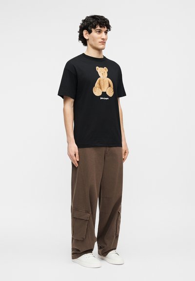 Junger Mann steht und trägt ein schwarzes T-Shirt mit einem Teddybär-Motiv, braune Cargo-Hosen und weiße Turnschuhe vor einem einfarbigen Hintergrund.