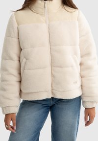 Veste matelassée couleur crème avec un tissu texturé et duveteux. Dotée d'un col montant, d'une fermeture éclair frontale et de panneaux cousus pour un look structuré.