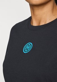 Musta puuvillapaita, jossa on pyöreä kaula-aukko ja turkoosi logo keskellä rintaa. Sileä pinta, istuva malli, lyhyet hihat.