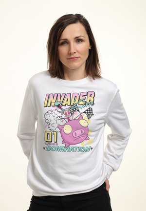 Kvinde med brunt hår iført en hvid sweatshirt med en farverig tegneseriegris, kapflag og tekst om verdensherredømme.