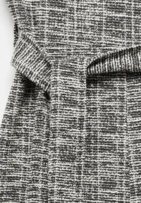 Tissu texturé noir et blanc avec un motif tissé, montrant un détail de boucle de ceinture pliée sur un vêtement.