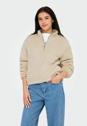 Femme aux longs cheveux foncés portant un pull en polaire beige zippé à moitié et un jean bleu taille haute, debout avec une main dans la poche.