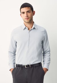 Világoskék hosszú ujjú ing kis geometriai mintával, gombos elől, nyitott gallérral, fekete nadrággal és övvel viselve.