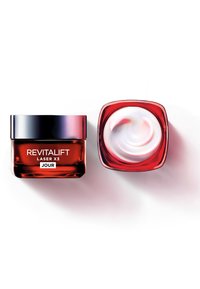 Pot de soin en verre rouge avec un couvercle noir. Contient un tourbillon de crème. L'étiquette indique "REVITALIFT LASER X3 JOUR" en police blanche sur le devant.