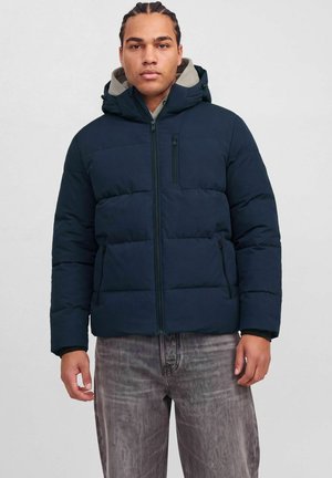 Homme aux cheveux tressés portant une doudoune bleu marine avec capuche, debout devant un fond clair uni.