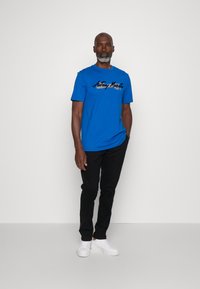 T-shirt in cotone blu con logo in testo nero, abbinata a pantaloni neri e sneakers bianche. Sfondo semplice senza ulteriori dettagli.