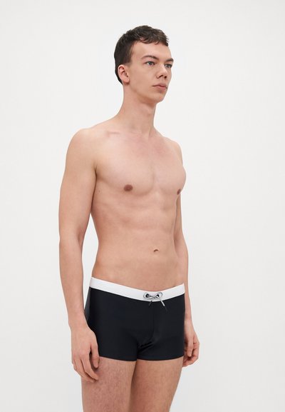 Shorts de bain noirs avec une ceinture élastique blanche et un cordon de serrage. Tissu doux et extensible, design ajusté et longueur au milieu de la cuisse.