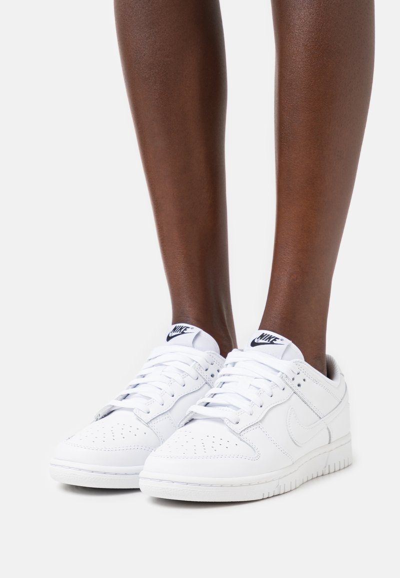 Baskets de sport blanches avec une boîte à orteils perforée, des lacets et un col rembourré. Présente un petit logo noir de Nike sur la languette et sur le côté.