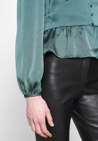Blouse satin teal avec de longues manches bouffantes et un ourlet froncé, associée à un pantalon en cuir noir ajusté. Détails de boutons sur le devant.