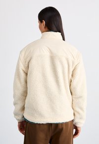 Giacca in pile color crema con un esterno testurizzato e morbido e un colletto beige liscio. Presenta una vestibilità rilassata e polsini elastici.